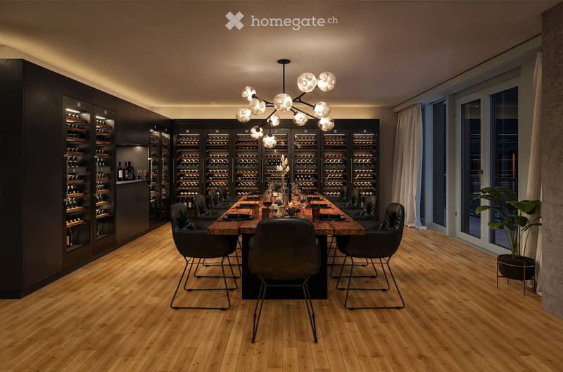 Large dining room with a wooden table and black chairs, surrounded by floor-to-ceiling wine racks. The room features a modern chandelier and a plant in the corner. The floor is hardwood and the walls are dark, creating a cozy and sophisticated atmosphere.