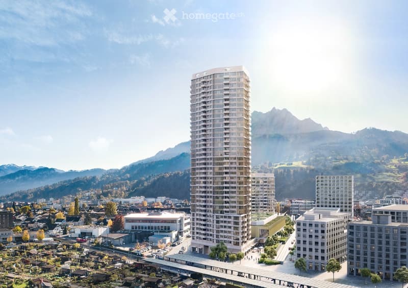 Tall residential high-rise building with multiple floors, surrounded by other buildings and mountains in the background.