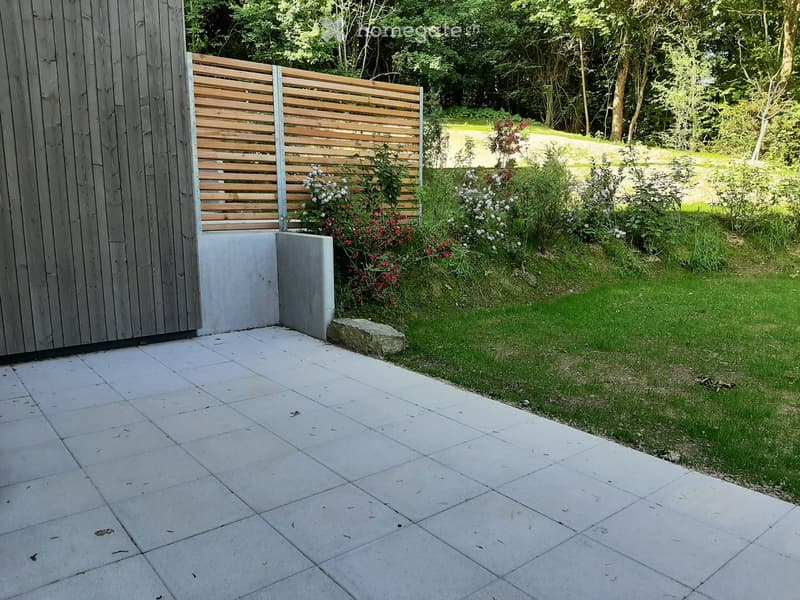Garden area with a paved patio, wooden fence, and lush greenery including flowering plants and shrubs.