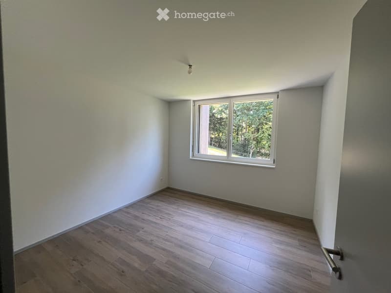 Empty room with light-colored walls, wooden flooring, and a single window showing greenery outside.