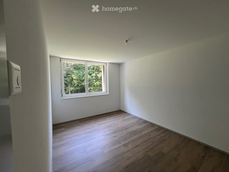 Empty room with light-colored wooden flooring and white walls, featuring a single window with white frames and a small electrical panel on the left wall.
