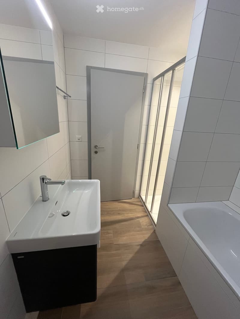 Modern bathroom with white tiles, a white sink with a chrome faucet, a bathtub, and a large mirror. The floor is wooden, and there is a glass-enclosed shower area visible in the background.