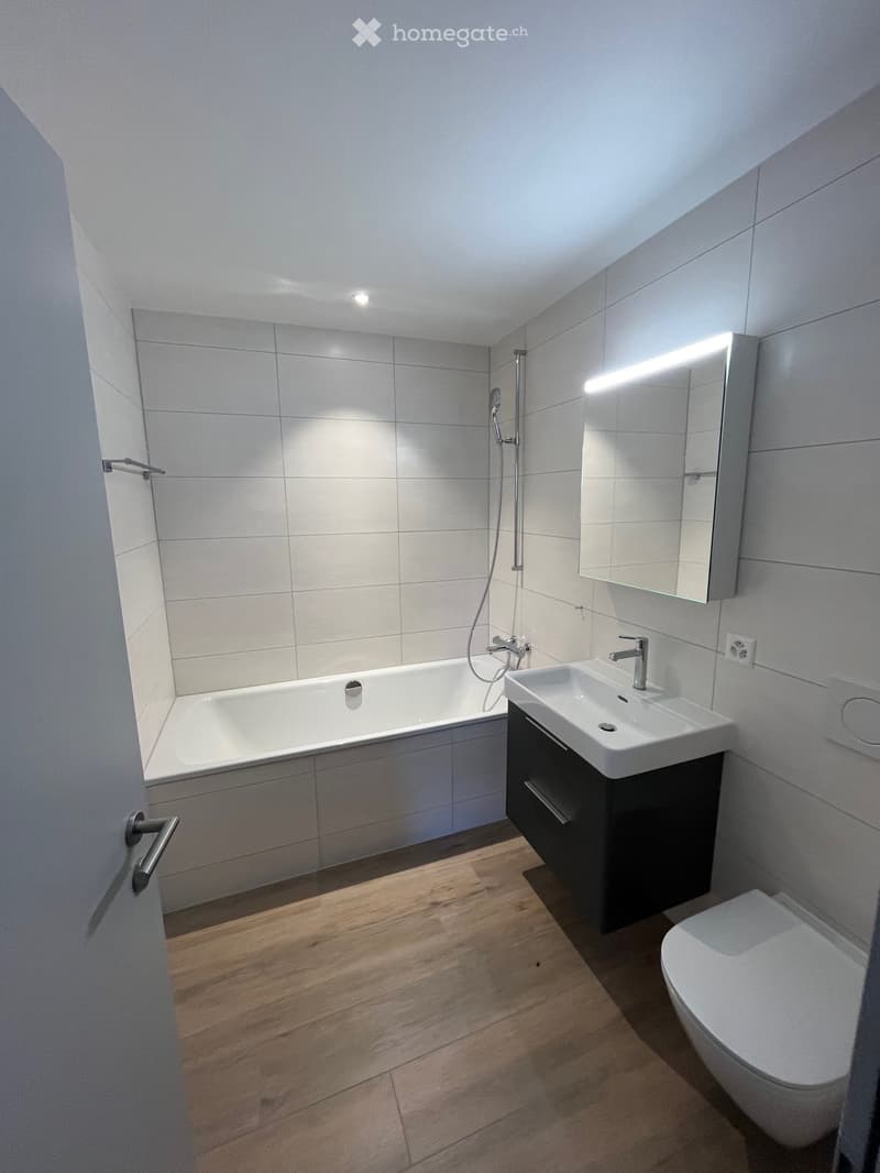 Modern bathroom with a bathtub, sink, toilet, and a large mirror with built-in lighting. The walls are tiled, and the floor is wooden.