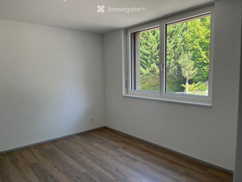 Empty room with light grey walls, wooden flooring, and large windows showing greenery outside.