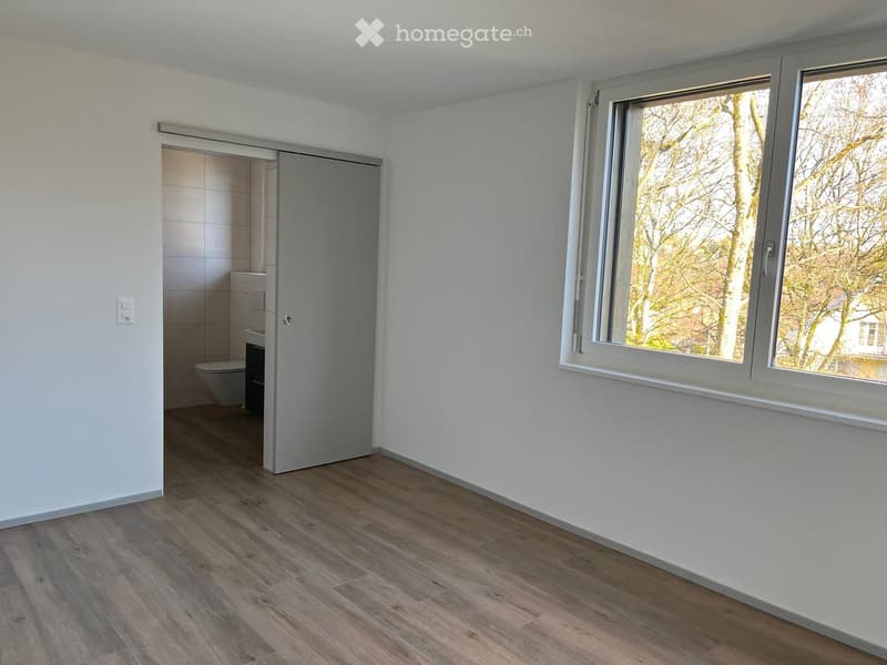 An empty room with white walls, wooden floor, sliding door leading to a bathroom, and a large window