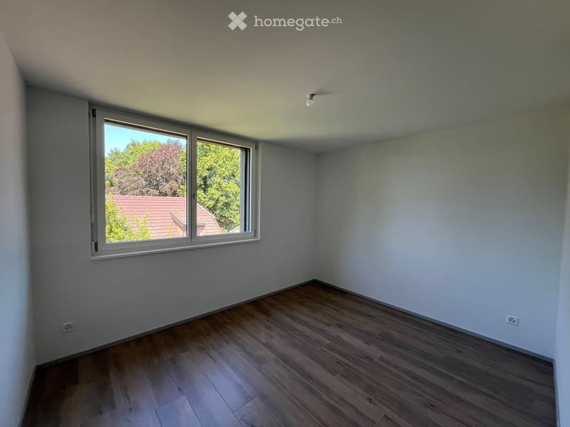 Empty room with light grey walls, wooden flooring, and a large window with a view of trees and a neighboring roof.