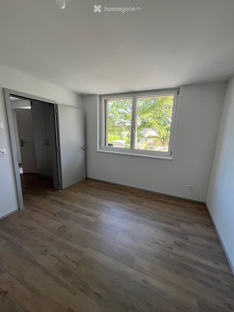 Empty room with light grey walls, wooden flooring, and a large window with a view of greenery outside. The room has a door leading to another space.