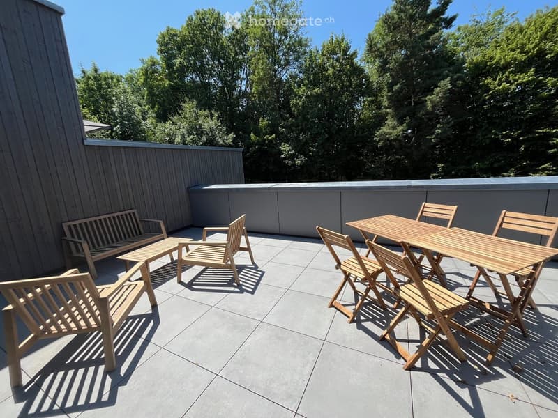 Wooden table and chairs, along with a bench, are arranged on a tiled balcony with a wooden railing and a view of trees.
