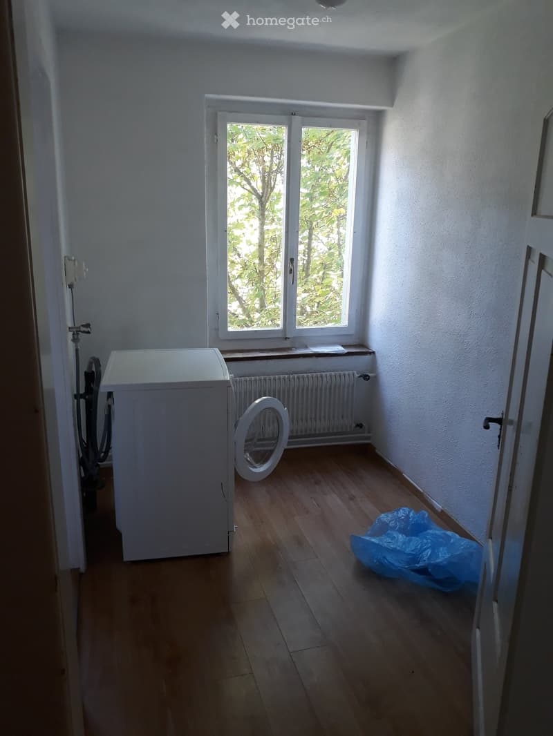 room with wooden floor, white walls, window with white radiator, white washing machine, blue plastic bag