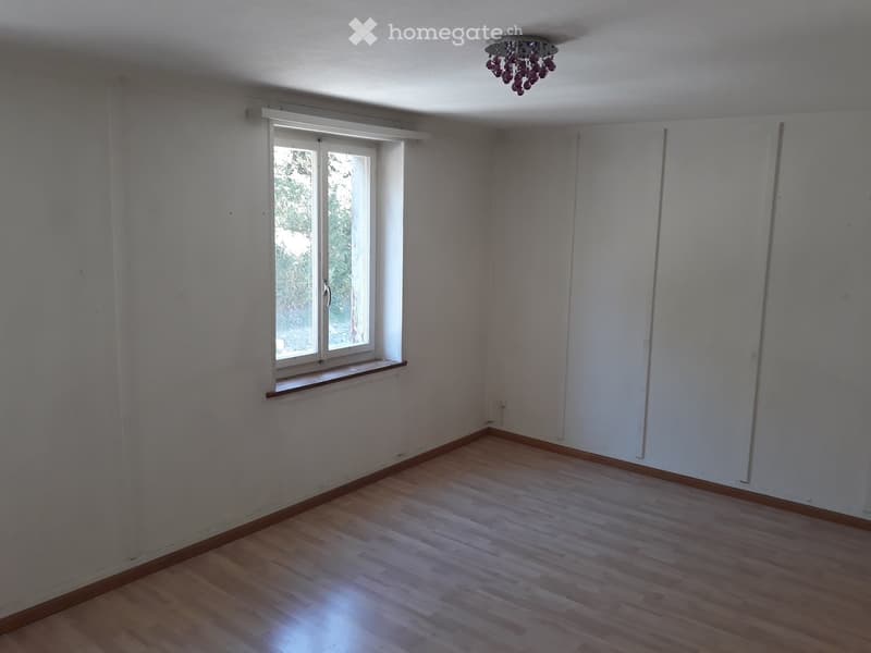 Empty room with wooden floors, a window, white walls, and a chandelier.