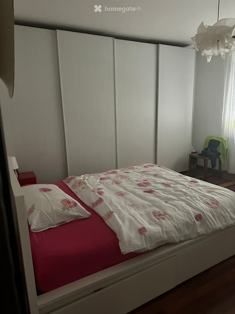 Bedroom with a double bed, white walls, white bed frame, red bed sheets, floral bedspread, white pillow, white nightstand, white lamp, white sliding wardrobes, wooden floor, white ceiling lamp, small wooden table, green cloth, chair, white curtains