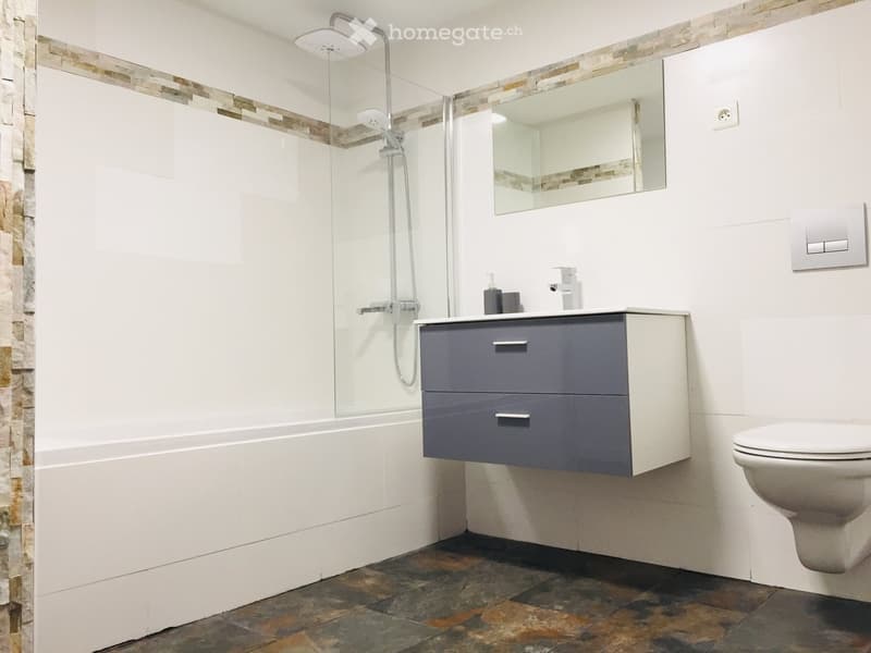 White tiles, shower with glass door, mirror, white vanity, sink, soap dispenser, toilet, tile floor
