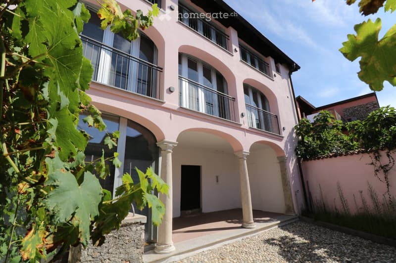 A two-story pink building with arched entrances, large windows, and balconies, surrounded by greenery and grapevines.