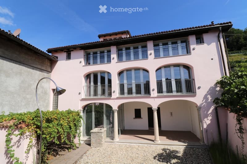 Two-story pink house with large windows, balconies, and a covered entrance area with columns. The house features a stone pathway and surrounding greenery.