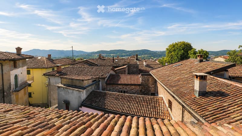 Roof tops of a village with terracotta tiles, chimneys, and distant mountains under a clear blue sky.