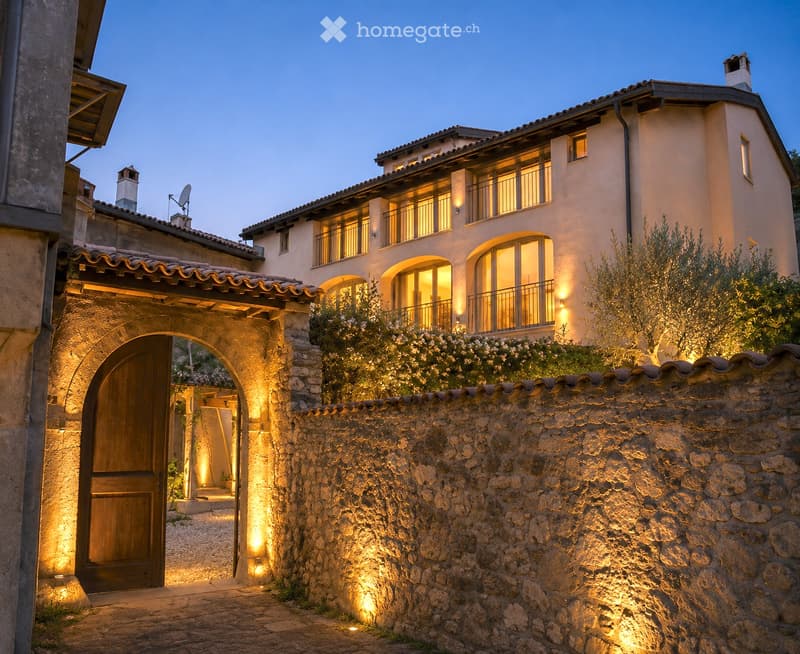 A two-story house with a stone wall entrance, arched wooden door, and illuminated windows. The building features a tiled roof and multiple balconies.