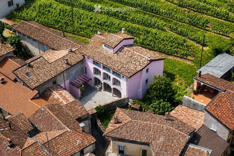 Aerial view of a large, pink villa with terracotta-tiled roofs, surrounded by vineyards and greenery. The villa features multiple wings, balconies, and a courtyard.