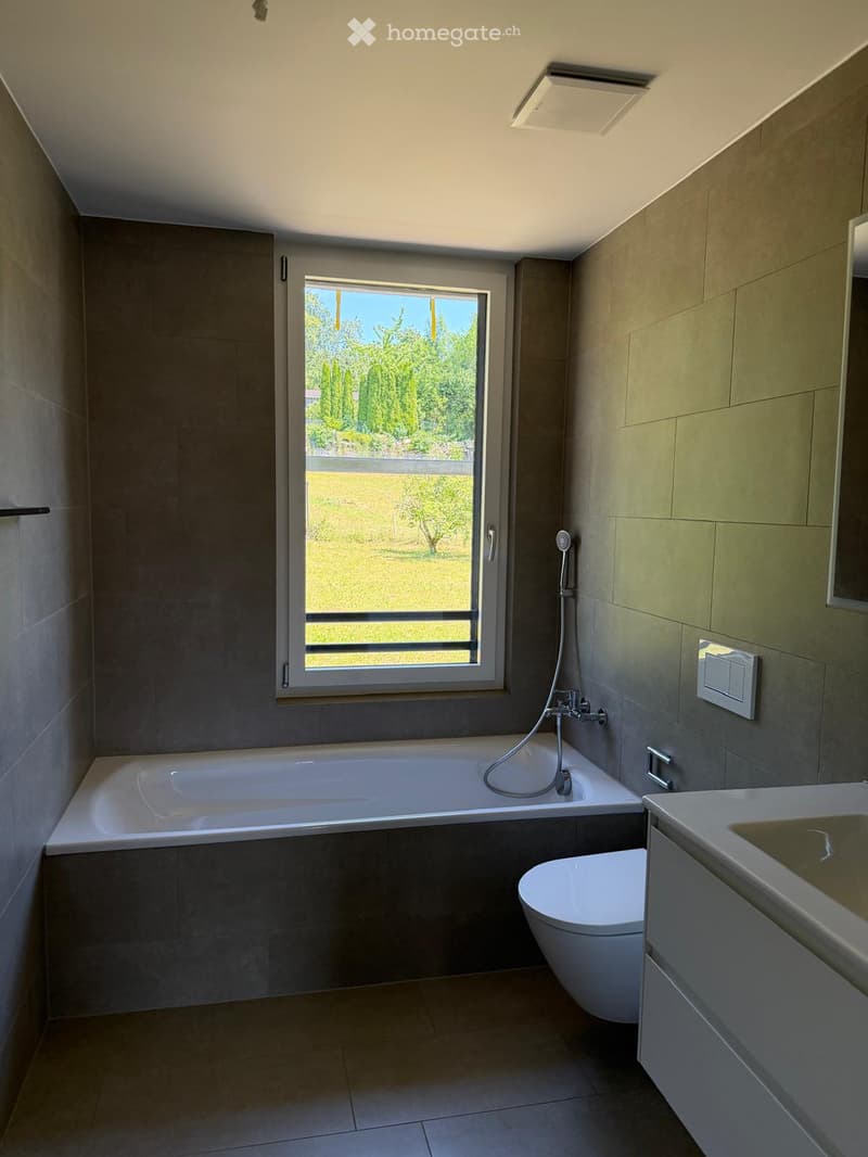 Modern bathroom with a bathtub, toilet, sink, and showerhead. Large window offers a view of greenery outside.