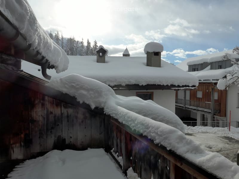 Wooden building with snow covered roof, wooden balcony railing, wooden wall with dark stains, windows covered with glass.