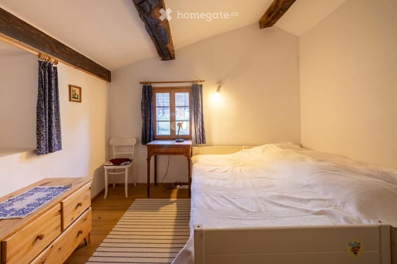 small bedroom with bed, wooden dresser, chair, desk, exposed wooden beams, small window with blue curtains