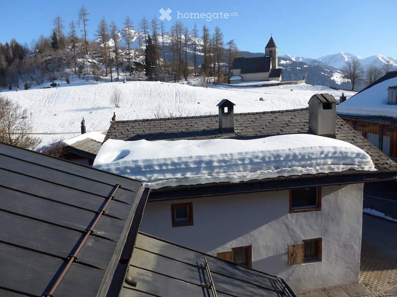 A snow-covered roof, black slate tiles, windows, wooden doors, mountains in the background, a small chapel, snow on the ground, pine trees.