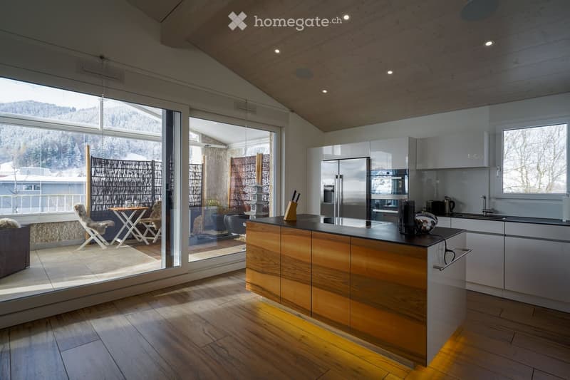 Open plan living room and kitchen, modern design, wooden flooring, large windows, glass doors leading to a balcony, wooden kitchen island, stainless steel appliances, view of mountains and trees