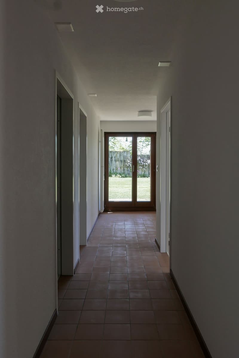 A long, narrow hallway with tiled flooring and white walls, leading to a glass door that opens to a garden area.