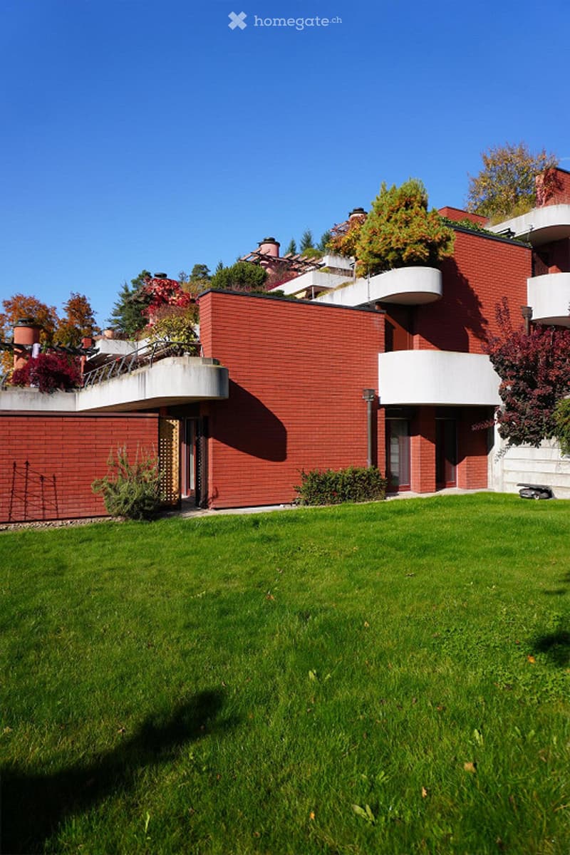 The house is multistory, featuring red brick exterior walls, white concrete balconies, and a green grassy front yard. Several trees and plants are visible around the building, with one tree growing on the wall.