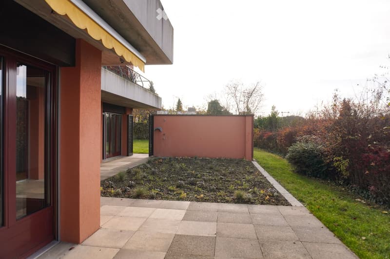 Garden with grass, red entrance door, building wall with glass windows, walkway