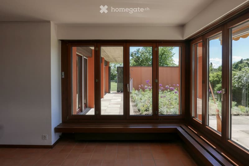 Empty room with large wooden-framed windows, tiled floor, and a wooden bench along the wall. The view shows a garden with flowers and greenery.