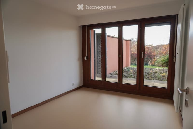 An empty room with wooden sliding doors, a white wall, and an open balcony on the outside. There is a power outlet on the wall and a wooden baseboard trim.