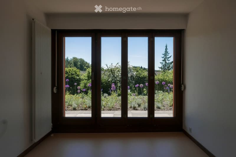 Empty room with large sliding glass doors opening to a garden, light-colored walls, and a tiled floor.