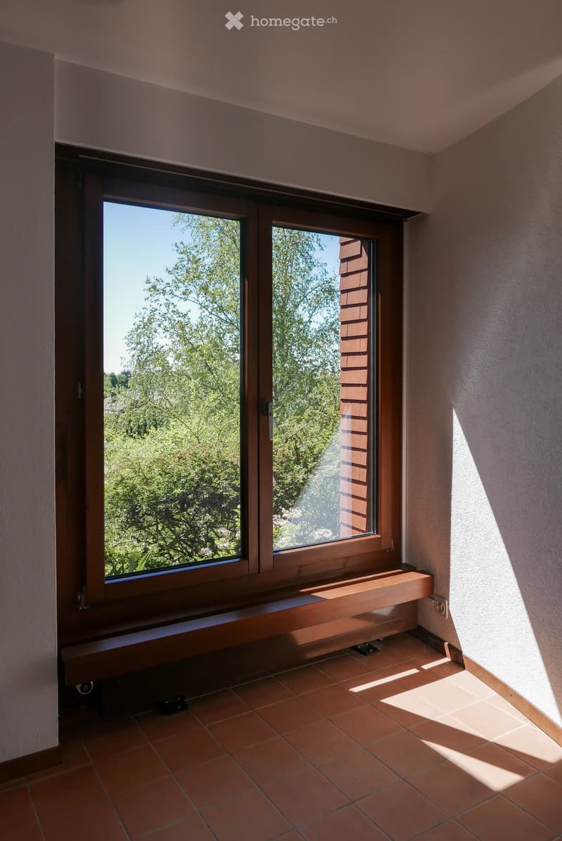 Empty room with terracotta tiled floor, large window with wooden frame, and a wooden bench beneath the window. Sunlight streams in, casting shadows on the floor.