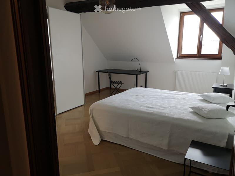Large wooden bed, white bedspread, desk with lamp, wooden floor, white walls, small folding chair, nightstands with lamps, radiator heater.
