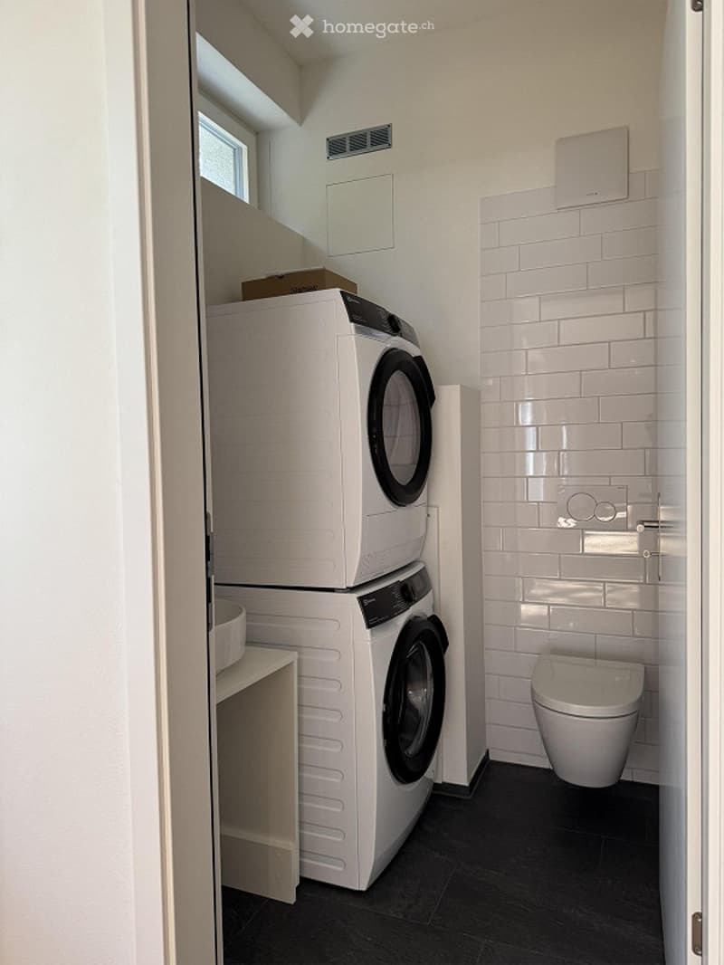 a modern compact laundry room with white tiled wall, floor and ceiling, two stacked washing machines, a small table, and a toilet.