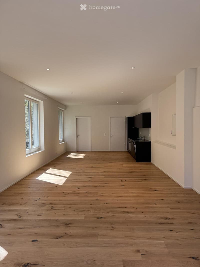 Empty room with wooden floors, two doors, windows, and a kitchen unit