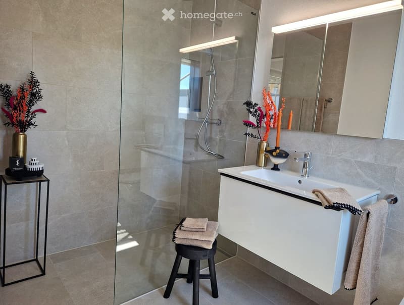 Modern bathroom with a white sink, large mirror, glass shower enclosure, decorative vase with flowers, and folded towels on a stool.