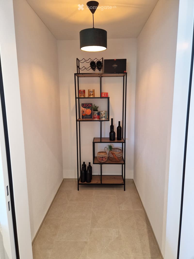 A narrow hallway with beige tiled flooring and white walls, featuring a modern black shelving unit with decorative items including bottles, books, and baskets.