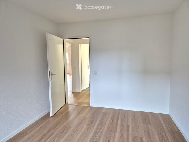 white walls, wooden floor, open door, interior with visible hallway and another room