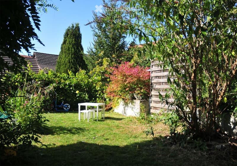 Small green garden with a white table and stool. Tall bushes and shrubs are present for privacy. The garden is mostly shaded.