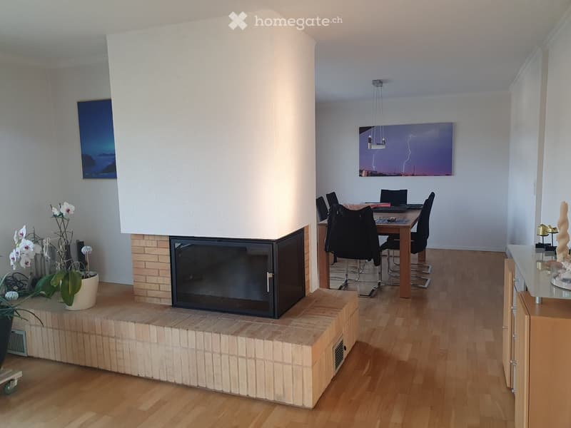 Living room with wooden floors, white walls, fireplace, dining table with 6 chairs, wooden desk, wall painting, and table lamp