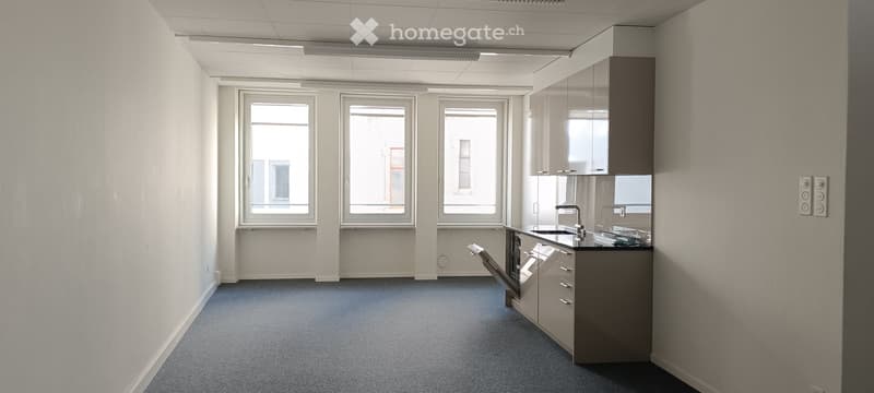 Empty room with white walls, gray floor, three windows, kitchen area with sink, cabinets, and dishwasher