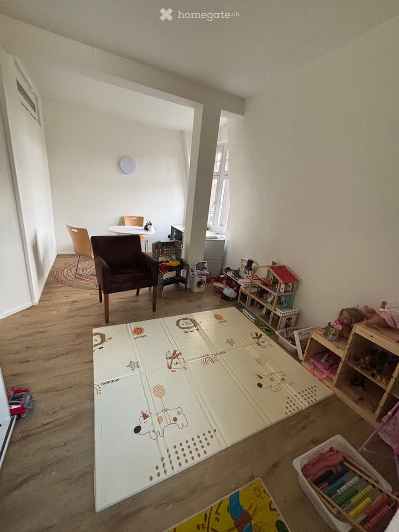 A playroom with a large playmat, a wooden chair, a small table, a white desk, shelves with toys, and a rug
