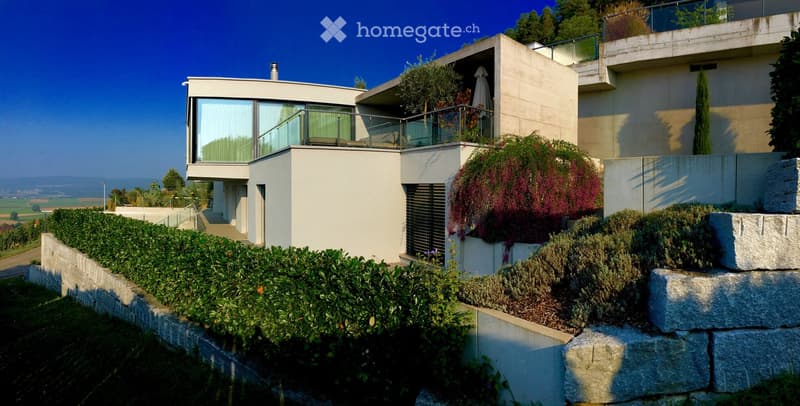 Modern house, concrete walls, glass windows, balconies, surrounded by stone walls and greenery