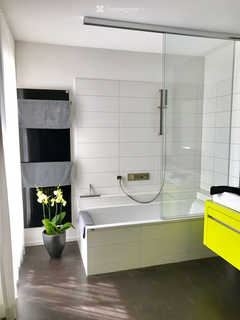 White bathroom with glass shower enclosure, bathtub, white and yellow cabinets, white tiles, potted flower, towel rack, silver fixtures