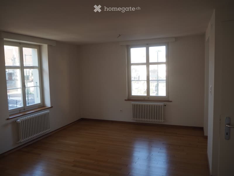 An empty room with white walls, wooden floors, and two windows with white frames. There are white radiators mounted on the walls. The room appears well-lit due to natural light coming through the windows. There is an open door on the right side of the roo