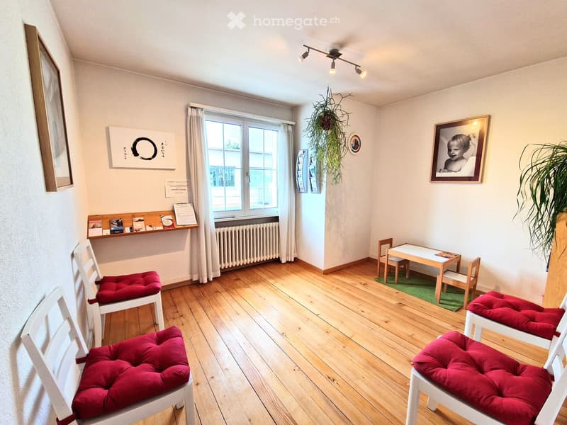 Room has hardwood floor, four red cushioned chairs, small table with green rug, small children's chairs and table, radiator, books and papers on shelf, pictures on wall
