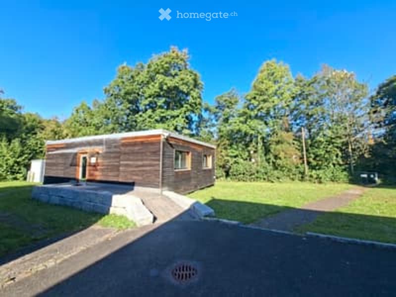A small wooden house with raised ground floor, surrounded by greenery and trees.