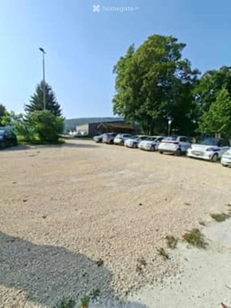 A parking lot with gravel surface and multiple cars parked, surrounded by greenery.