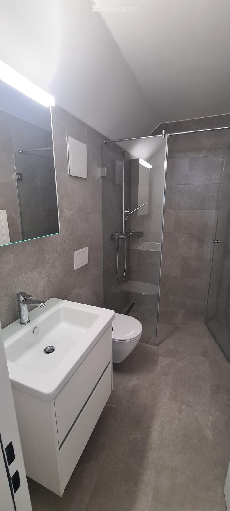 modern bathroom with white sink, grey tiles, walk-in shower with showerhead, toilet, mirror with light, tiled floor